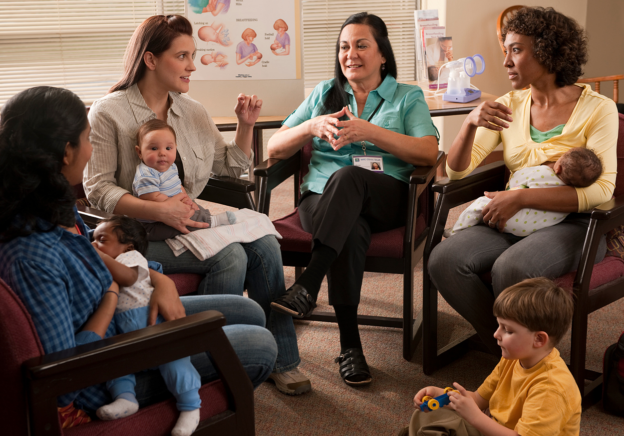 Women sitting in a circle for a lactation class. Mothers are holding their infants. There is a young child playing on the ground.